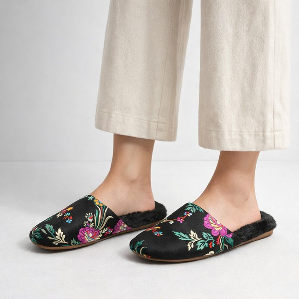 Black slippers with colorful floral patterns worn with beige pants on a light gray background