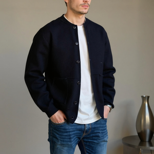 Man wearing a black jacket over a white shirt and blue jeans, standing against a neutral background.