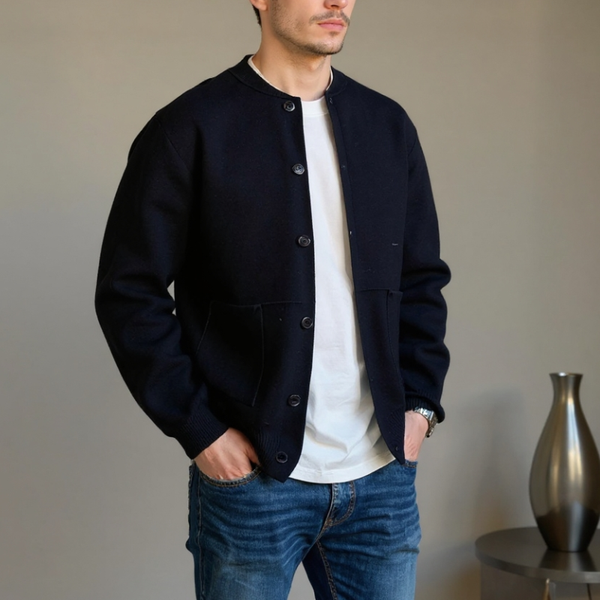 Man wearing a black jacket over a white shirt and blue jeans, standing against a neutral background.