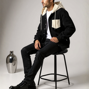 Man sitting on a stool wearing a black and beige jacket with a white shirt underneath, on a plain background.