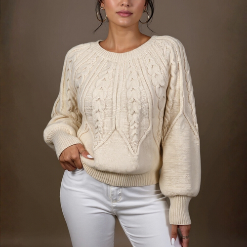 Woman wearing a cream cable knit sweater and white pants against a brown background