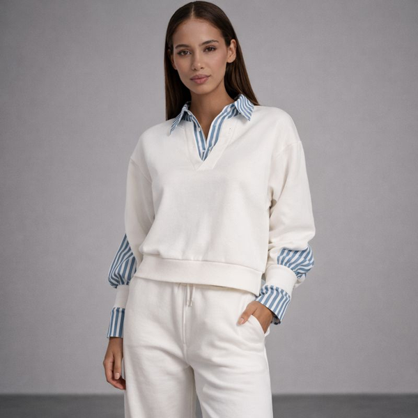 Woman wearing a white sweater with striped cuffs and collar over a blue and white striped shirt, paired with white pants on a gray background.