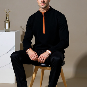 Man sitting on a stool wearing a black outfit with an orange zipper, with a neutral background.