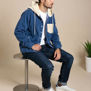 Man sitting on a stool wearing a blue jacket, white shirt, and jeans against a beige background.