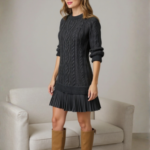 Woman wearing a black cable knit sweater dress with pleated skirt and brown knee-high boots in a neutral setting.