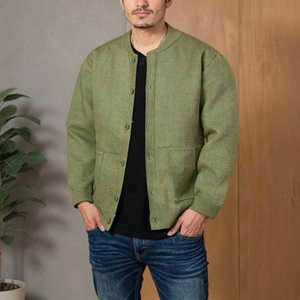 Man wearing a green jacket and blue jeans standing against a wooden wall.