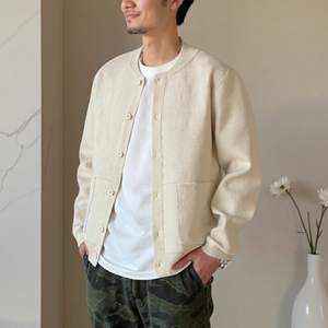 Man wearing a beige jacket over a white shirt with camouflage pants, standing against a neutral wall.
