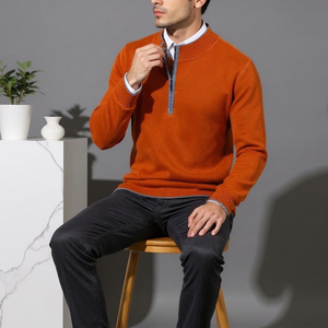 Man wearing an orange sweater sitting on a wooden stool against a gray background