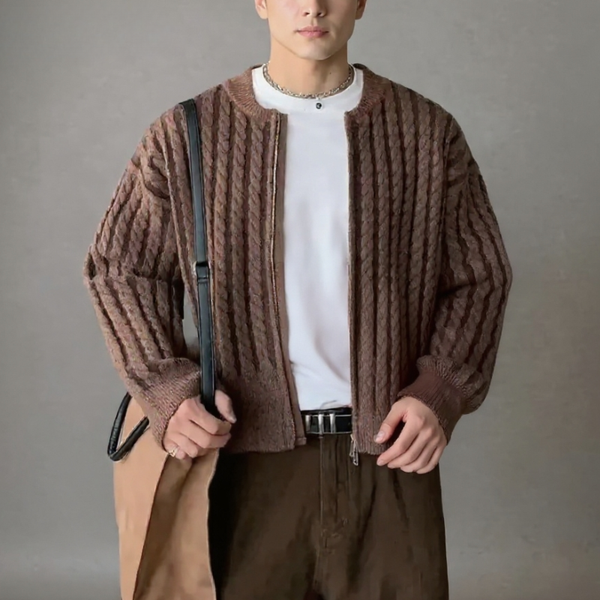 Person wearing a brown textured cardigan over a white shirt, holding a beige coat against a gray background.