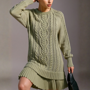 Person wearing a green cable knit sweater and pleated skirt against a neutral background