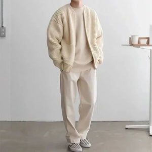 Person wearing a beige cardigan, sweater, and pants in a minimalistic room.