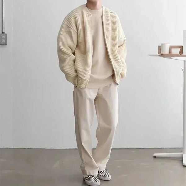 Person wearing a beige cardigan, sweater, and pants in a minimalistic room.