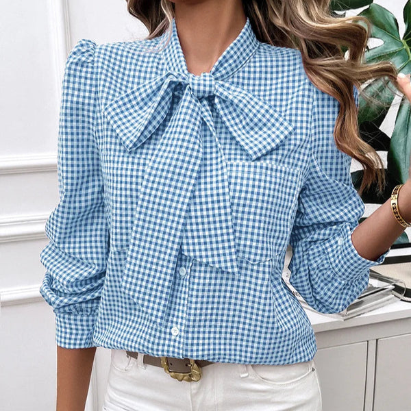 Woman wearing a blue checkered blouse with a bow tie and white pants in an indoor setting.