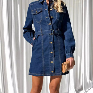 Person wearing a blue denim dress with a belt against a white curtain background