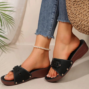 Black sandals with brown soles worn by a person with denim pants and a pearl bracelet.