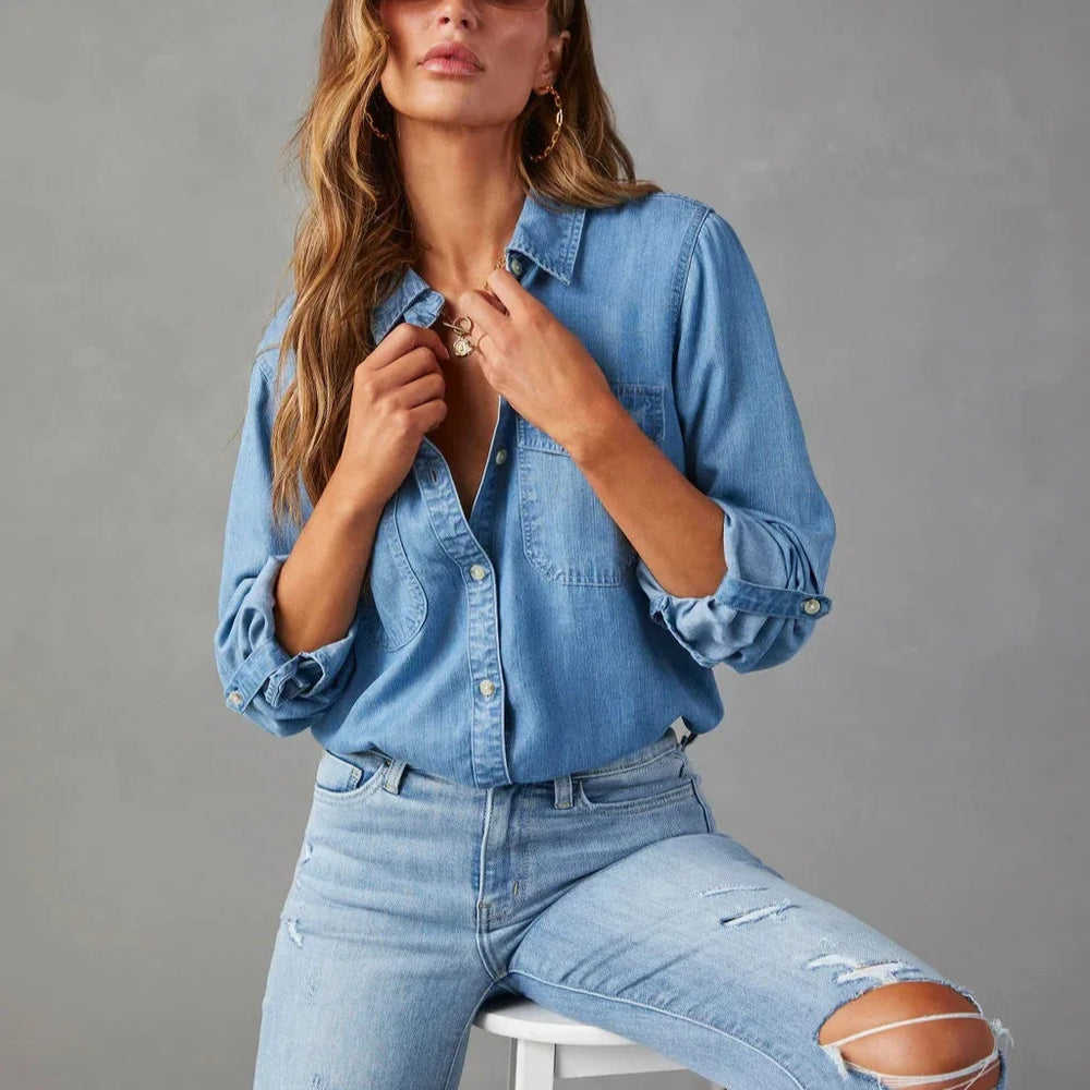 Woman wearing a light blue denim shirt and jeans, posing against a gray background.