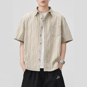 Person wearing a beige short-sleeve shirt over a white t-shirt with black pants on a light gray background