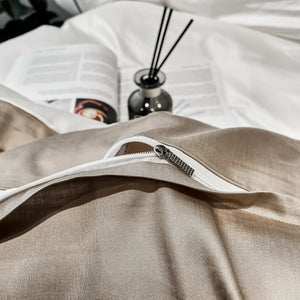 Beige pillow with a zipper on a white surface, with a diffuser and book in the background.