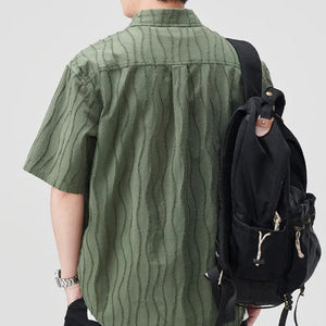 Person wearing a green shirt and black shorts with a backpack, standing against a light gray background.