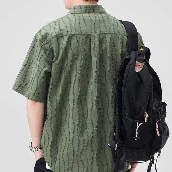 Person wearing a green shirt and black shorts with a backpack, standing against a light gray background.