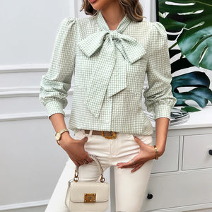 Woman wearing a green checkered blouse with a bow tie, holding a beige handbag.